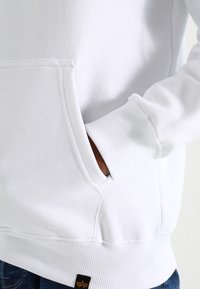 Witte hoodie met een voorzak, ribgebreide boorden en zoom. Opvallende stikdetails en een klein merkje aan de onderkant.