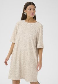 Bēšs īsām piedurknēm apģērbs, kas pārklāts ar teksturētiem spīguļiem, ar apaļu apkakli un brīvu piegriezumu. Gluda auduma ar spīdīgiem akcentiem.