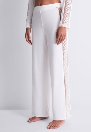 Femme portant un pantalon blanc taille haute à jambes larges avec des panneaux en dentelle transparente sur les côtés, debout pieds nus devant un fond uni.