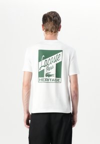 Baltos medvilnės marškinėliai, su žalia grafika ant nugaros, kurioje yra užrašas „Lacoste Paris Heritage“ ir krokodilo logotipas.