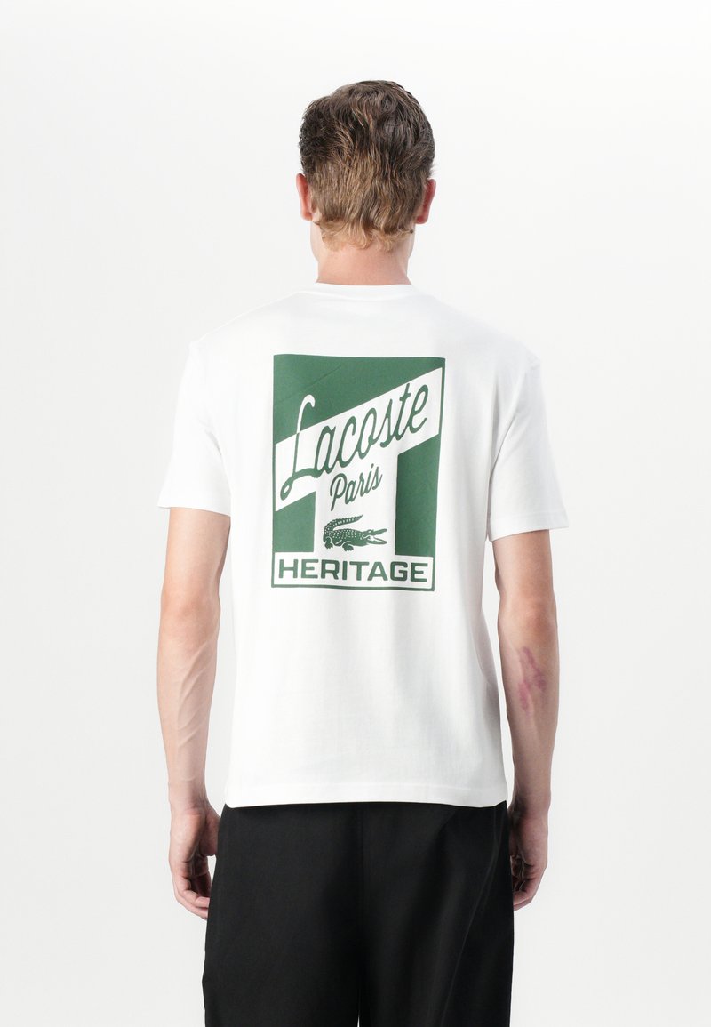 Baltos medvilnės marškinėliai, su žalia grafika ant nugaros, kurioje yra užrašas „Lacoste Paris Heritage“ ir krokodilo logotipas.