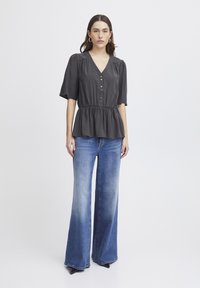 Graue, kurzärmlige Bluse mit V-Ausschnitt und Knopfleiste; geraffte Taille. Kombiniert mit weiten, blauen Jeans und schwarzen High Heels.