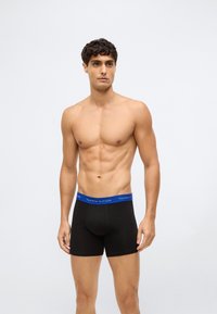 Homem em forma com cabelo curto e encaracolado, vestindo boxers pretos com uma cinta azul da Tommy Hilfiger, de pé contra um fundo claro e simples.