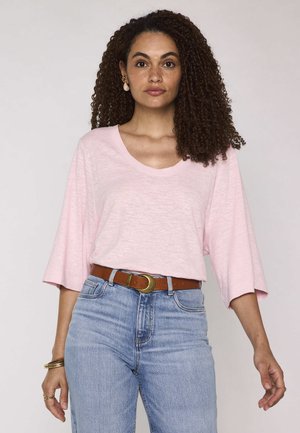 Donna con capelli ricci che indossa un top rosa chiaro a maniche larghe, jeans blu a vita alta, cintura marrone e orecchini e braccialetti dorati.