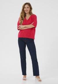 Un pull v-neck rose vif à manches courtes, en tricot texturé, associé à un jean droit bleu foncé et des sandales à talons blancs à lanières.