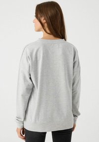 Sudadera gris con mangas largas, cuello redondo y puños acanalados. Textura suave, corte relajado, detalles de costura visibles, combinada con pantalones negros.