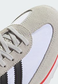 Grijze suède en witte mesh sneaker met zwarte strepen en rode accenten, ronde vorm en veters van gestructureerde stof.