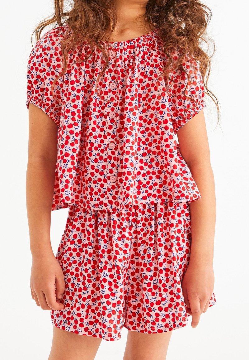 Jeune fille portant un chemisier à manches courtes rouge et blanc à motifs cerises et un short assorti sur fond blanc.
