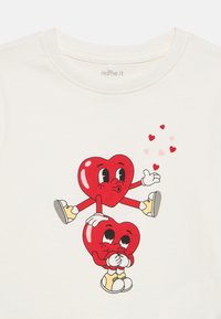 Κρεμ μπλουζάκι με εκτύπωση δύο καρτούν καρδιών: η μία καθιστή, η άλλη όρθια με τα χέρια ανοιχτά, περιτριγυρισμένες από μικρές κόκκινες καρδιές.