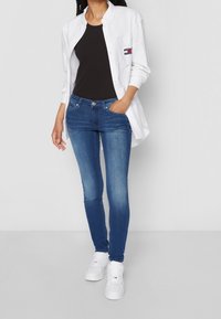 Vit skjorta med knappar över en svart tanktop, kombinerad med åtsittande blå jeans och vita sneakers. Synliga fickdetaljer på skjortan.