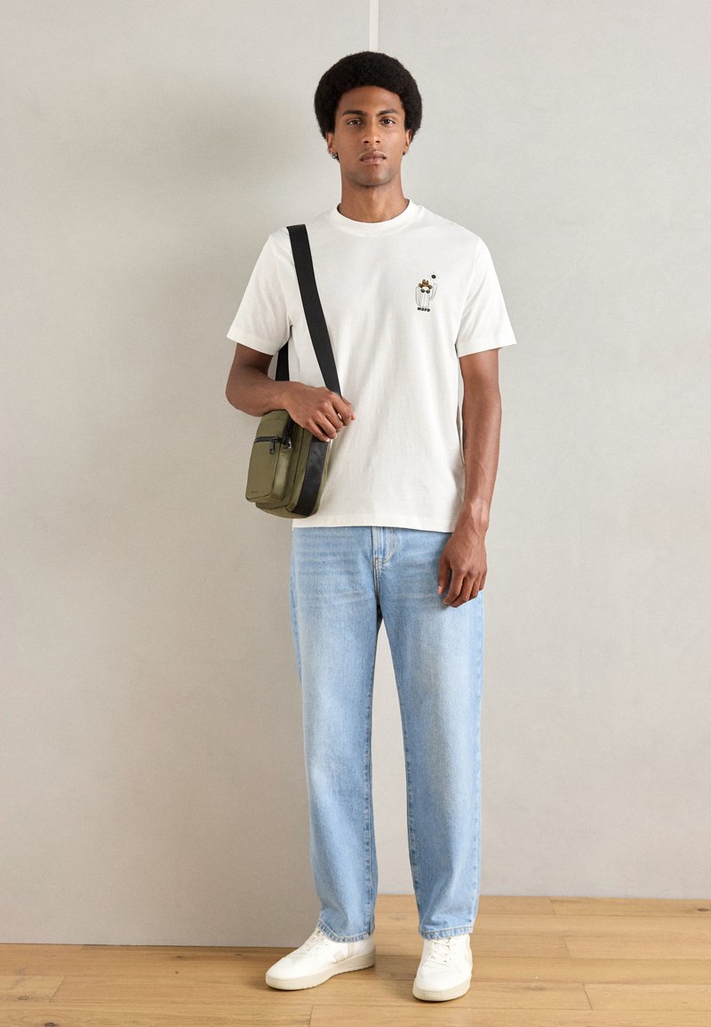 Weißes Baumwoll-T-Shirt mit einem kleinen Grafik, hellblaue Jeans und weiße Sneakers. Eine grüne Umhängetasche mit einem schwarzen Riemen rundet den Look ab.