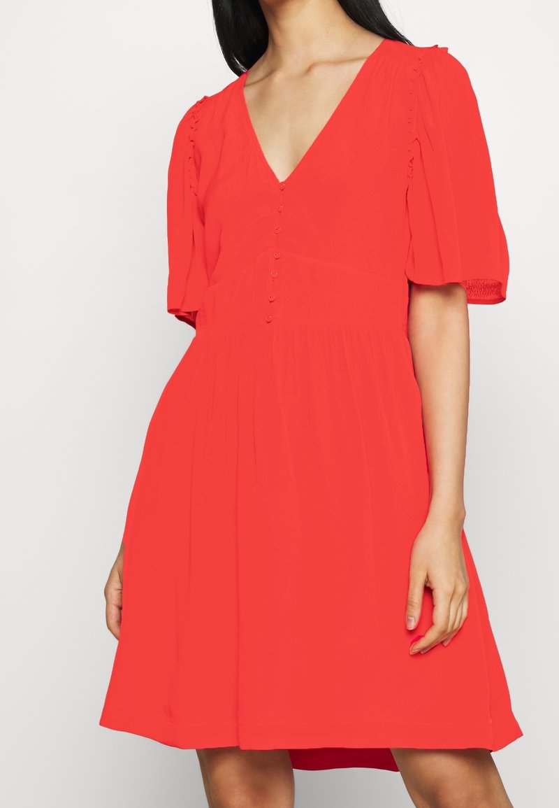 Rode jurk met een diepe V-hals, korte pofmouwen en een knoopsluiting aan de voorkant. De stof is lichtgewicht en textuurrijk, met een uitlopende rok.