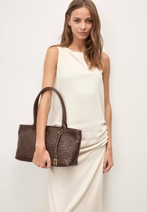 Žena u bijeloj haljini bez rukava drži smeđu pletenu kožnu torbu s dvostrukim ručkama i detaljem resica.