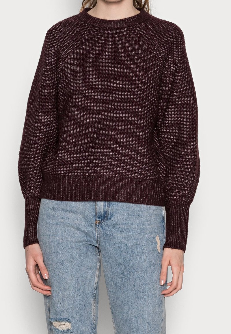 Pull en maille bordeaux avec une texture côtelée, encolure ronde et épaules tombantes, associé à un jean bleu clair délavé.