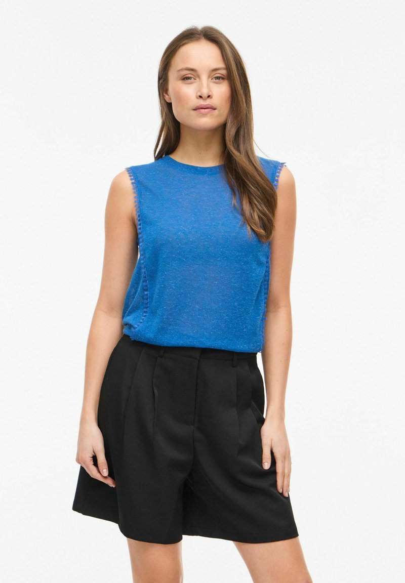 Femme aux longs cheveux bruns portant un haut bleu sans manches et un short noir taille haute, debout devant un fond blanc uni.