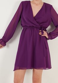 Žena v krátkých fialových šatech s průhlednými dlouhými rukávy a uvázaným páskem v pase, pózující s jednou rukou na bocích.