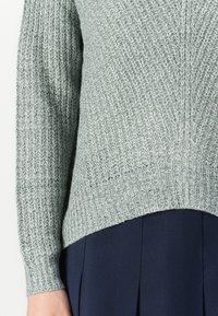 Lys grønn strikkegenser med teksturweave, ribbestrikkede mansjetter og kant. Har en lengre rygg enn fronten, kombinert med en marineblå skjørt.