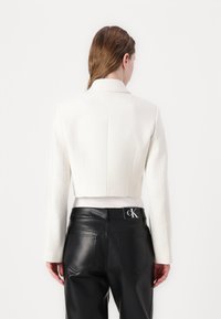 Veste blanche cropped avec manches longues, tissu texturé et un petit col. Portée par-dessus un pantalon en cuir noir avec un logo sur la taille.