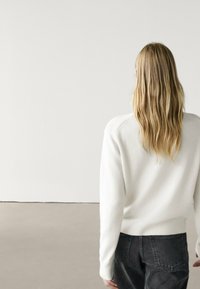 Valkoinen neulepaita, jossa on pyöreä pääntie ja ribbireunus, yhdistettynä tummaankin farkkuihin. Mallin hiukset ovat pitkät ja laineikkaat, kuvattu takaa.