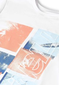 Biely textil s abstraktným dizajnom zloženým z nášiviek v pastelových farbách oranžovej, modrej a bielej, s čiastočne viditeľnými veľkými obkreslenými písmenami.