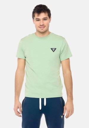 Jonge man draagt een lichtgroen T-shirt en donkerblauwe joggingbroek, staand tegen een effen witte achtergrond.