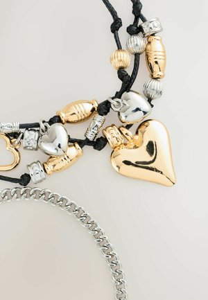 Zlaté a stříbrné přívěsky na černé šňůrce, včetně tvarů srdce a texturovaných korálků, se stříbrným řetízkem v popředí.