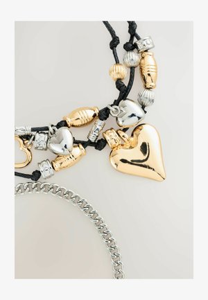 Zlaté a stříbrné přívěsky na černé šňůrce, včetně tvarů srdce a texturovaných korálků, se stříbrným řetízkem v popředí.