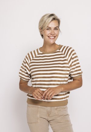 Lächelnde Frau mit kurzen blonden Haaren, die einen braun-weiß gestreiften Kurzarmpullover und beige Hosen trägt, vor einem schlichten Hintergrund stehend.