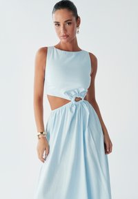 Une femme pose dans une robe découpée bleu clair avec un détail noué à la taille, accessoirisée de bracelets en or. Un maquillage subtil met en valeur son apparence.
