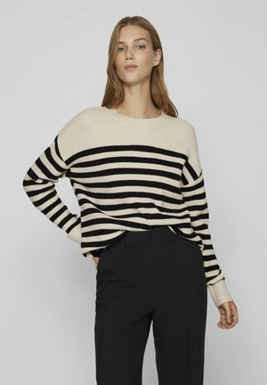 Suéter a rayas en crema y negro, con amplias rayas horizontales, puños acanalados y un corte relajado, combinado con pantalones negros.
