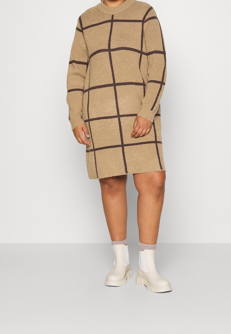 Personne portant une robe-pull beige à motif quadrillé foncé et des bottines blanches avec des chaussettes gris clair sur un fond uni.