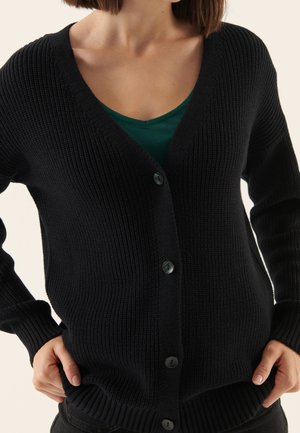 Femme portant un cardigan noir côtelé boutonné par-dessus un haut vert foncé à col en V, les mains légèrement glissées dans les poches d'un pantalon noir.