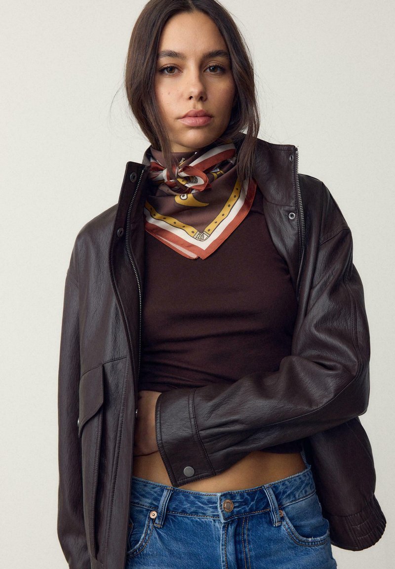 Femme portant une veste en cuir marron foncé, un haut raccourci marron, un jean bleu et un foulard en soie à motifs autour du cou sur un fond uni.
