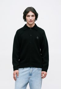 Cardigan negru tricotat cu guler, închidere cu butoane și tiv ribatit. Prezintă un mic logo brodat pe piept.
