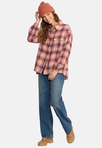 Rutigt flanellskjorta i rosa och orange nyanser, knäppning fram, med en bröstficka; ihop med lösa blå jeans och bruna skor.