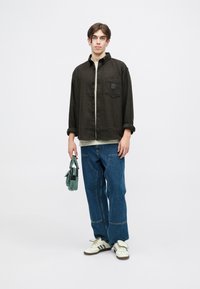 Jachetă căptușită cu carouri maro peste un tricou verde deschis, purtat cu blugi largi de culoare albastră și adidași albi cu dungi negre. Ține o geantă de culoare teal.