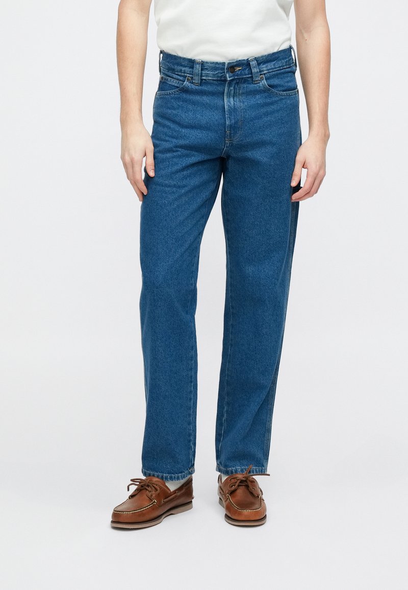 Jean en denim bleu foncé avec une coupe droite, cinq poches, et une fermeture à boutons à l'avant. Porté avec des chaussures bateau marron.