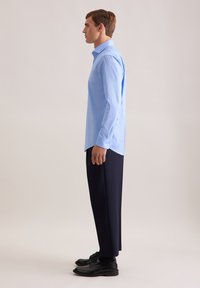 Camicia button-up a maniche lunghe di colore azzurro chiaro con colletto, abbinata a pantaloni navy e scarpe nere, che si trova davanti a uno sfondo neutro.