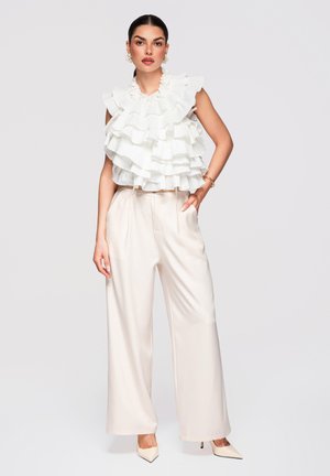 Kobieta w białym, warstwowym, falbaniastym topie bez rękawów, szerokich kremowych spodniach i szpilkach z zaostrzonym noskiem, stoi z jedną ręką w kieszeni.