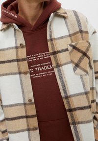 Camicia di flanella a quadri beige e bianca con una felpa marrone sotto. La flanella presenta bottoni marroni e una tasca sul petto. Texture morbida.
