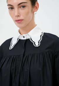 Blusa nera con colletto bianco ricamato dal design scallop. Presenta una texture plissettata e una chiusura con bottoni.