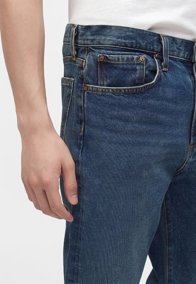 Gros plan sur la main d’une personne reposant à côté d’un jean en denim bleu avec poche avant visible et coutures des passants de ceinture apparentes.