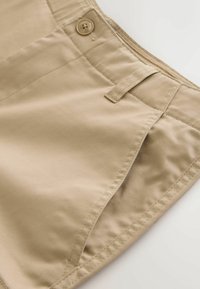 Gros plan sur l'avant d'un pantalon en coton beige montrant le bouton, la passants de ceinture et le détail de la poche fendue.