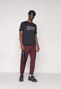 Melnā Nike T-kreklā ar logo, vīnu krāsas šaurās bikses, melni sporta apavi un melna ūdens pudele. Gluda tekstūra, brīvs piegriezums un sportisks dizains.