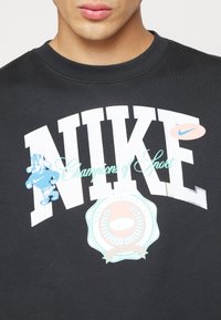 Persona con sudadera negra de cuello redondo con grandes letras blancas "NIKE", perro de dibujos animados azul, texto "Campeones del Deporte" y emblema de ropa deportiva en el pecho.