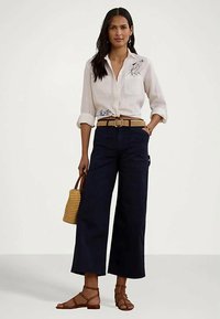 Ženska, oblečena v belo s cvetličnim vezenjem, mornarsko široke hlače, rjavi pleteni pas, rjave sandale z naramnicami in drži pleteno torbico.