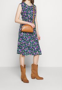 Kwiatowa sukienka z dopasowanym gorsetem i rozszerzaną spódnicą w kolorach zielonym, fioletowym i białym; połączona z brązową skórzaną torebką na ramię i beżowymi kowbojkami.