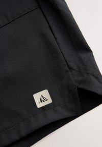 Tissu noir avec section perforée, couture détaillée et petit logo réfléchissant carré avec un motif triangulaire.