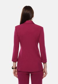 Blazer magenta aderente con collo a punta, maniche a tre quarti e polsini arrotolati. Tessuto liscio con silhouette su misura e senza chiusure visibili.