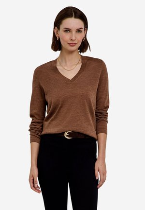 Femme aux cheveux courts et bruns portant un pull marron à col en V, un pantalon noir avec ceinture, de petites boucles d'oreilles dorées en anneau et un fin collier doré.